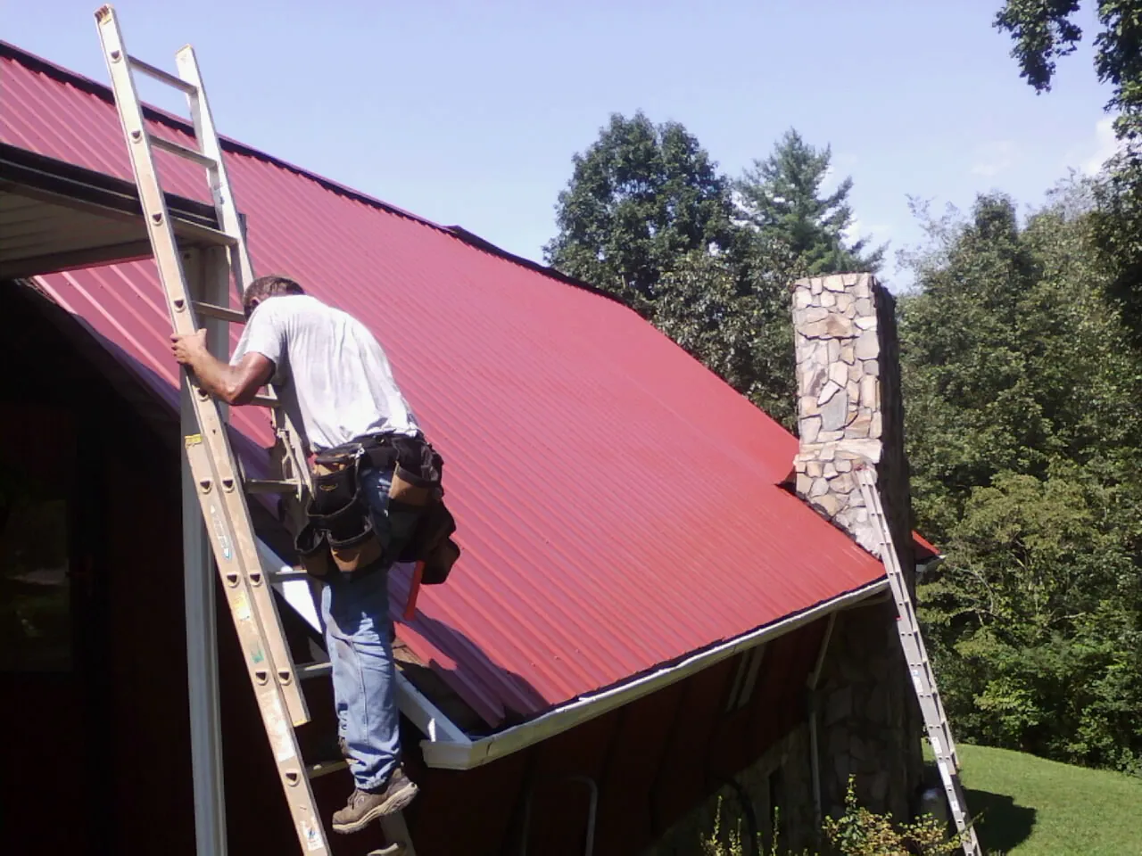 Roofer on a ladder beside a red metal roof and stone chimney — Soffit & Fascia Repair by Roofing Brothers in Safety Harbor